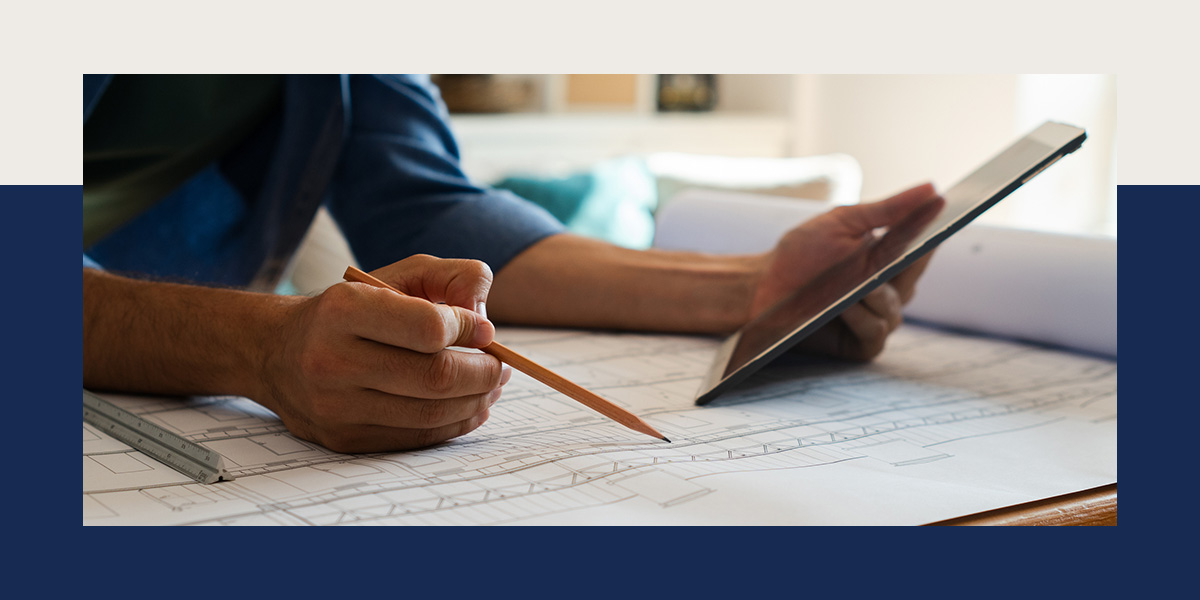 Close-up of a person's hands holding a pencil and a tablet while reviewing architectural blueprints on a table, with a ruler nearby.
